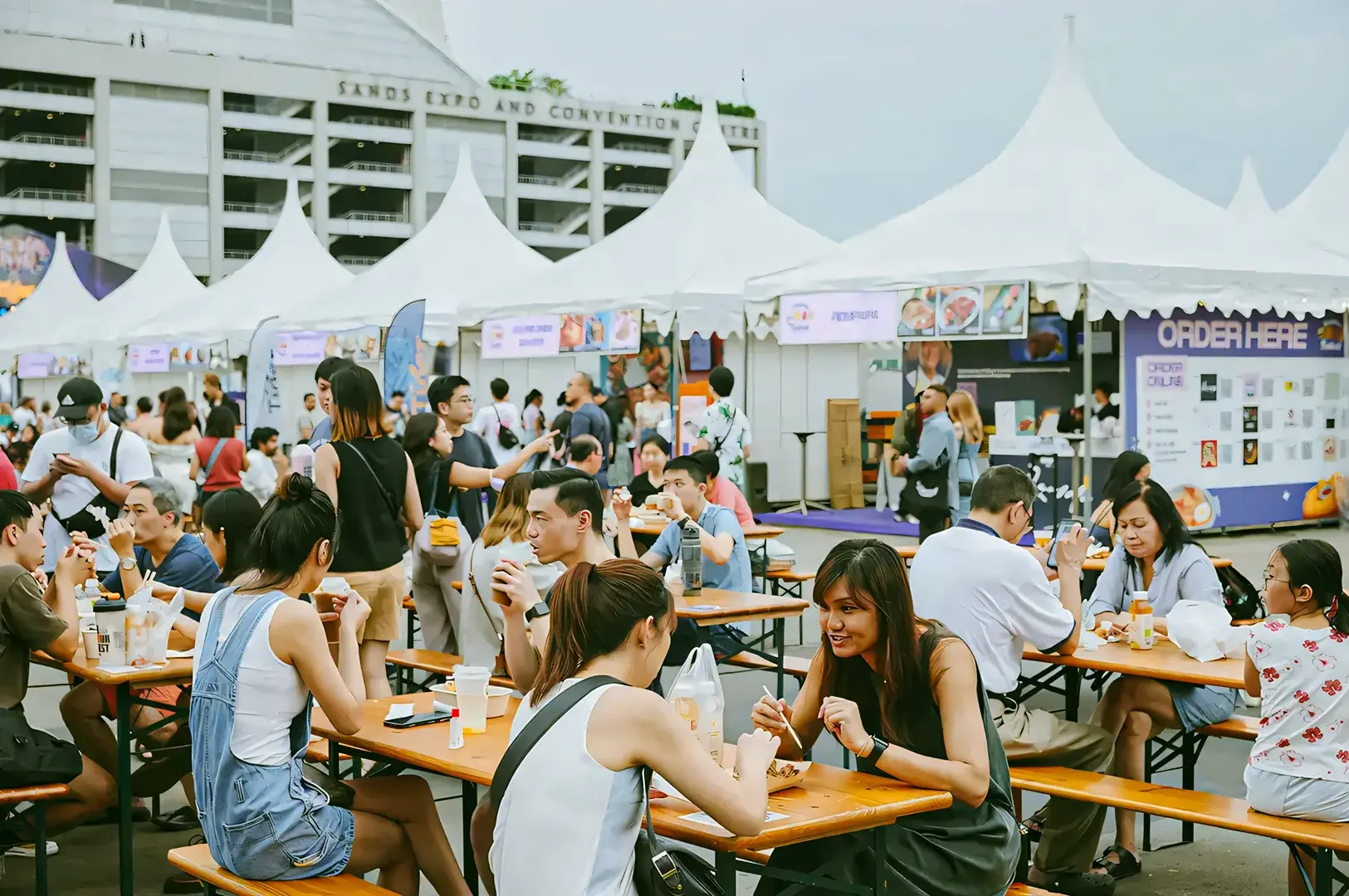 Outdoor Singapore food festival with white canopy tents, stalls offering various dishes, and crowds dining at long wooden communal tables.