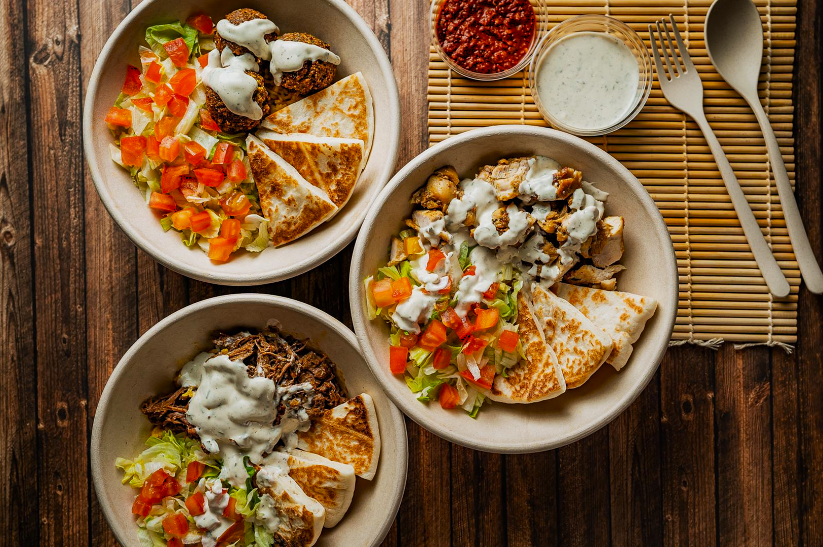 Three bowls of vibrant salads with diced tomatoes, lettuce, grilled meat, and quesadilla slices, drizzled with creamy dressing. Fork and spoon nearby.