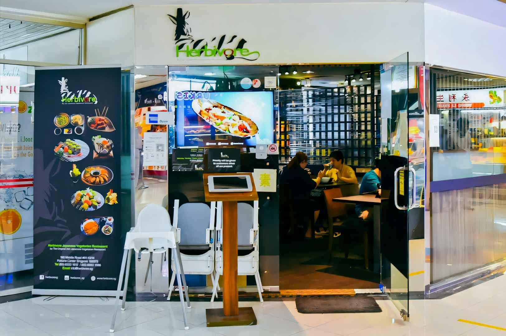 Glass‑front entrance of Herbivore vegetarian restaurant in Fortune Centre, featuring menu posters, digital food displays, and indoor dining tables.
