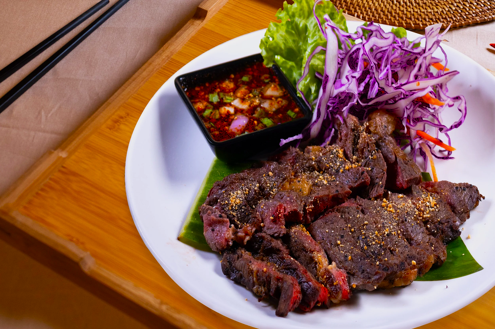 Grilled steak slices on a white plate with purple cabbage, lettuce, and a small bowl of dipping sauce. Set on a wooden board with chopsticks.