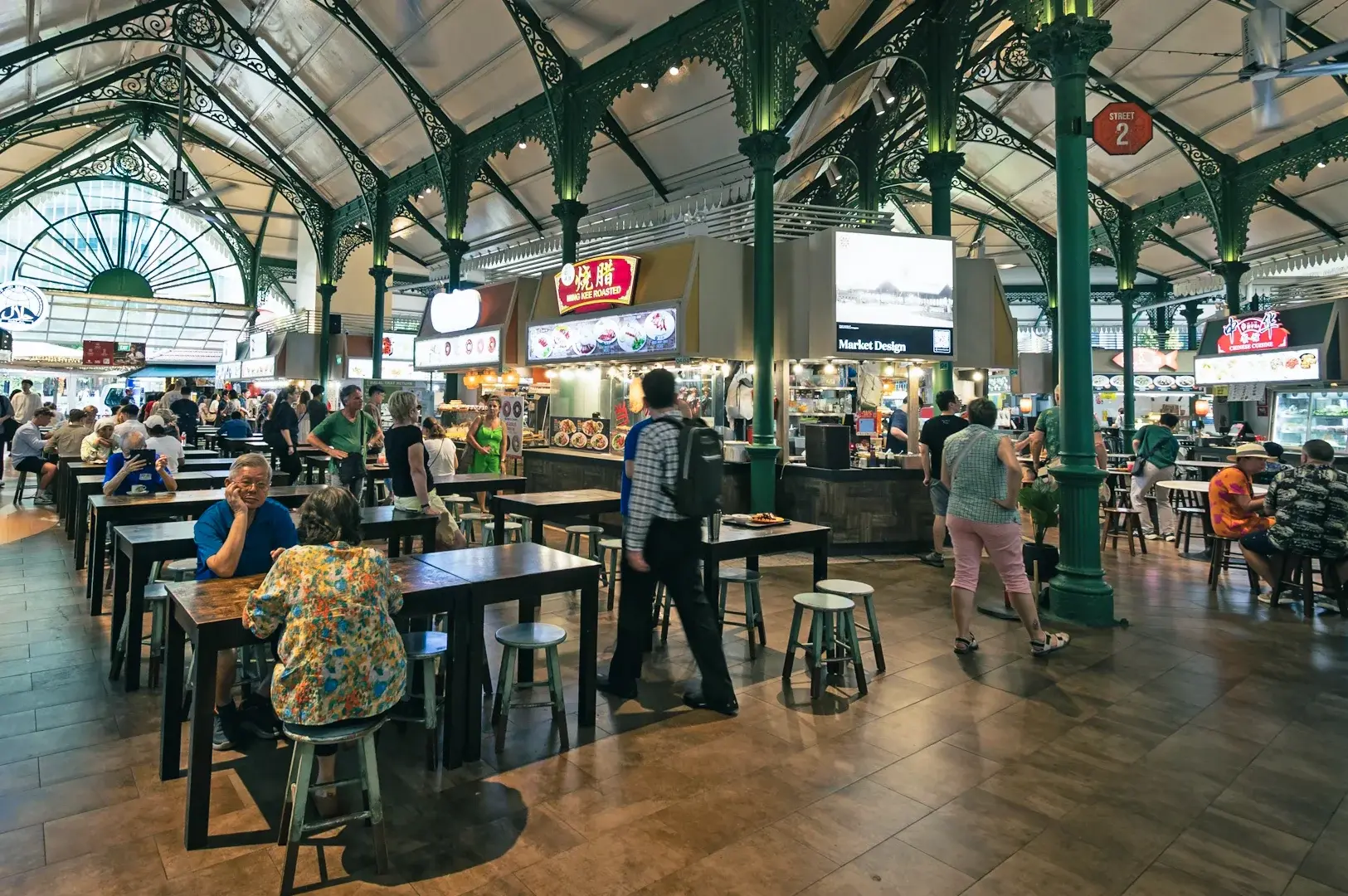 Interior of a Singapore hawker center featuring multiple food stalls, dining tables, and market-style architecture.