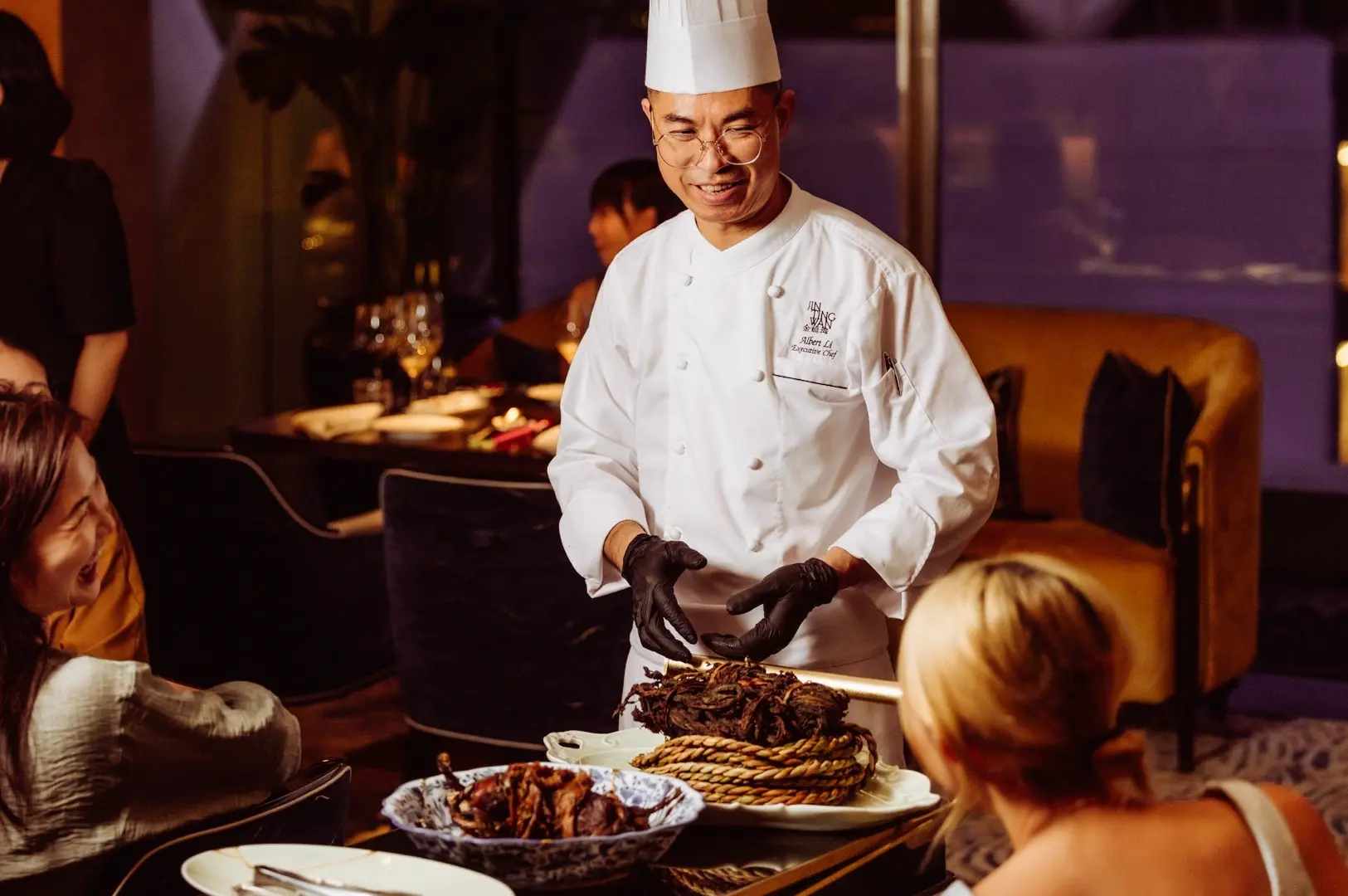 Chef in a white uniform and tall chef’s hat presenting assorted roasted meats tableside in an upscale restaurant, with diners seated around the service area.