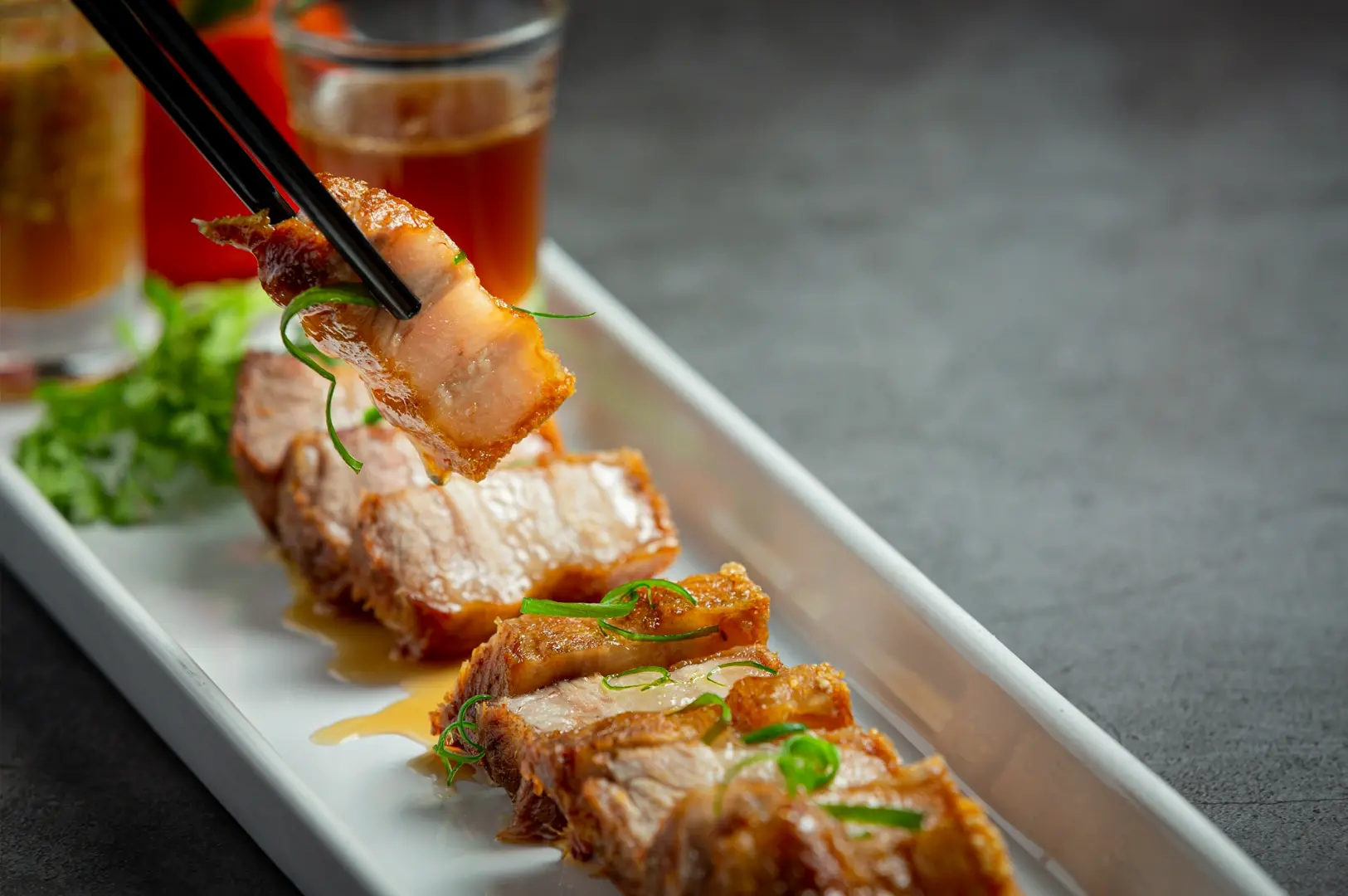 Chopsticks lifting a piece of crispy roasted pork belly garnished with green onions, served on a rectangular plate with dipping sauce and fresh greens in a fine Chinese dining setting.