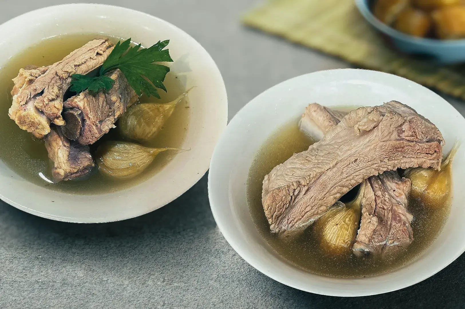 Two bowls of Bak Kut Teh pork rib soup with garlic cloves in a clear herbal broth.