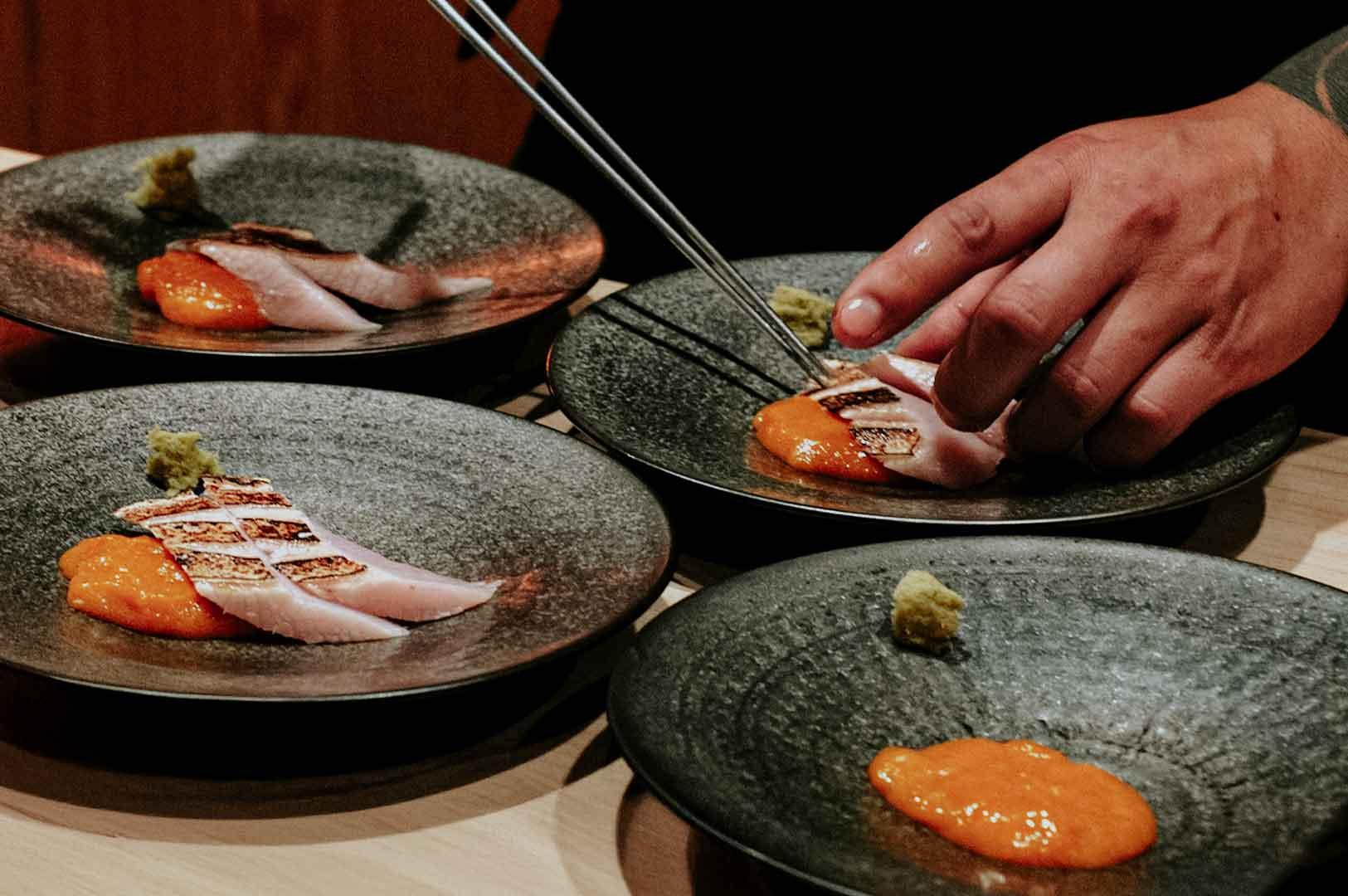 Chef carefully plating seared fish slices with bright orange sauce on textured ceramic plates at a Japanese omakase restaurant.
