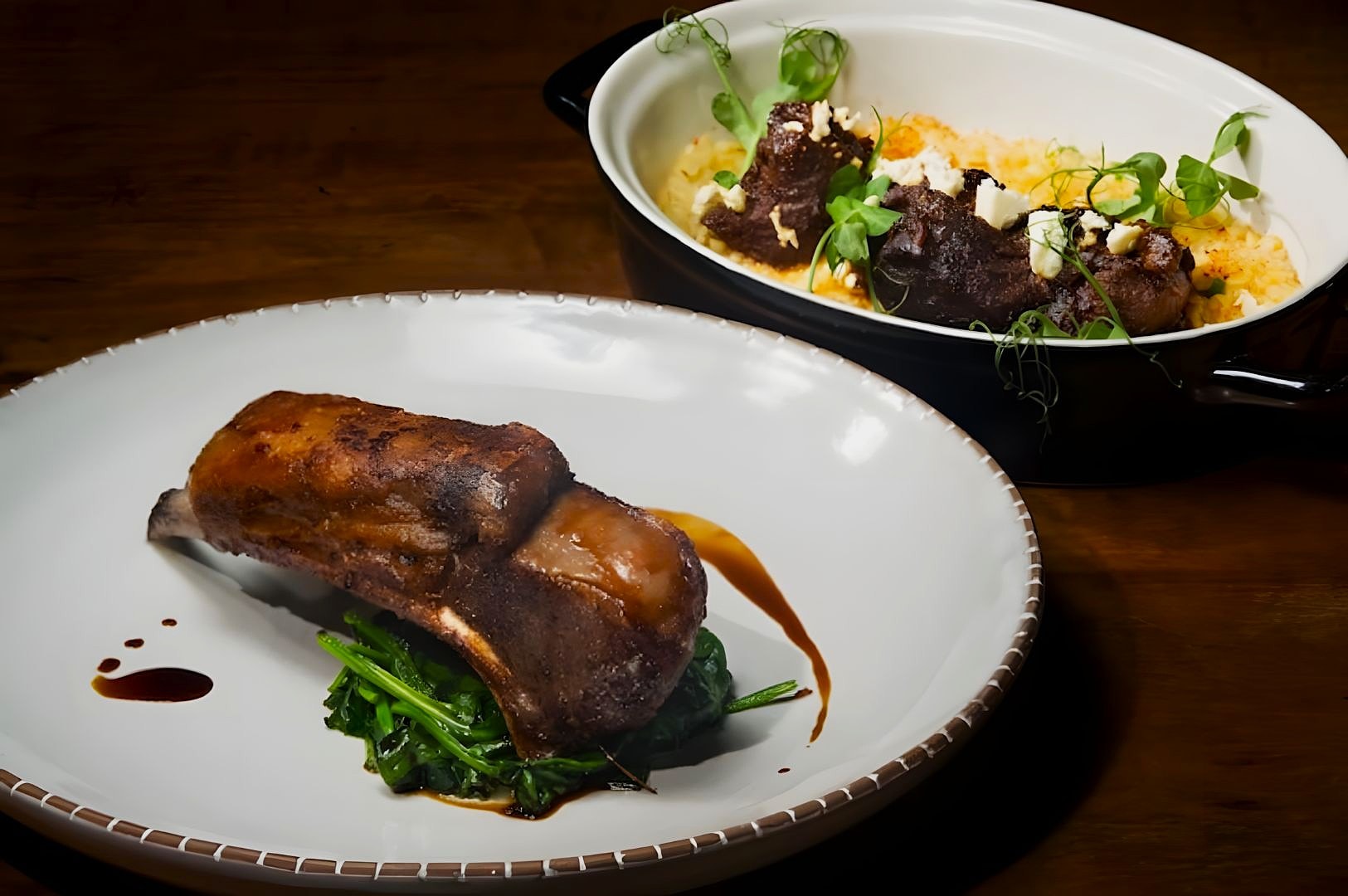 Roasted meat on sautéed greens with a dark sauce on a white plate; a casserole in the background contains meat with cheese and herbs.