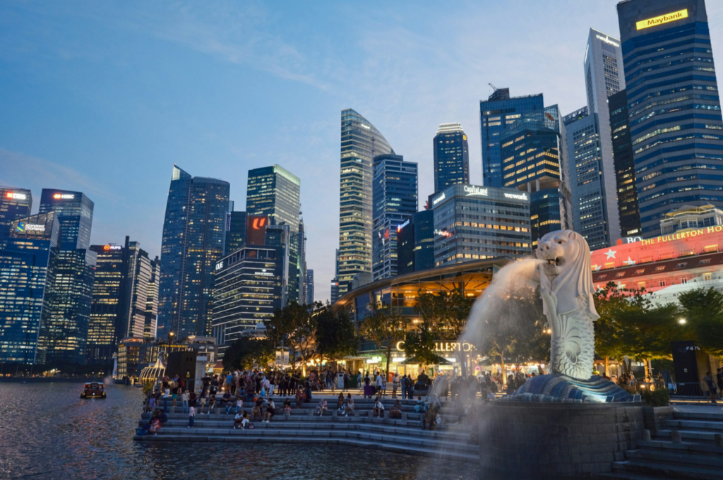 View of Singapore's skyline at dusk, featuring the iconic Merlion statue spouting water in the foreground, with illuminated skyscrapers in the background.
