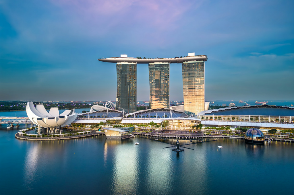 “Aerial view of Marina Bay Sands in Singapore at dusk, with its three illuminated towers and rooftop platform. The sky is a soft blue and pink.”