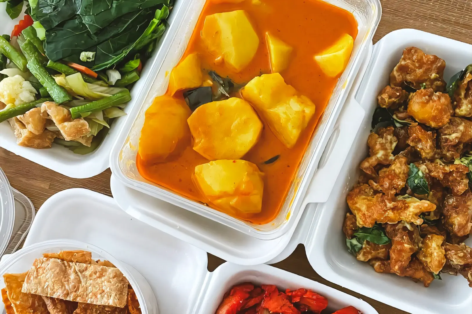 Top‑down shot of Singapore takeaway food in plastic containers featuring curry chicken, vegetables, fried items, and rice on a table.