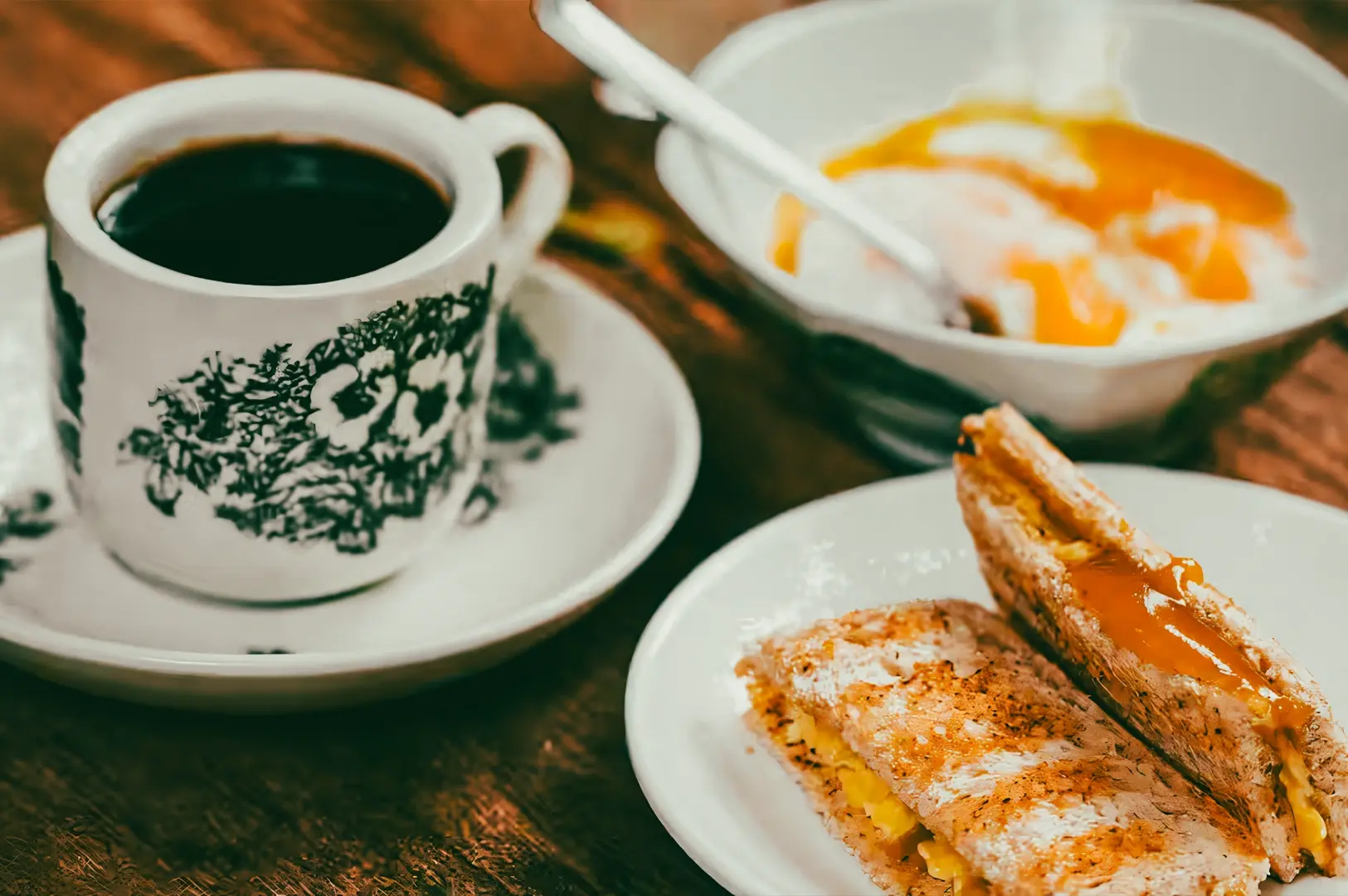 Medium close-up eye-level shot of traditional Singapore kopi served in a floral ceramic cup with kaya toast slices and soft‑boiled eggs on a wooden café table.