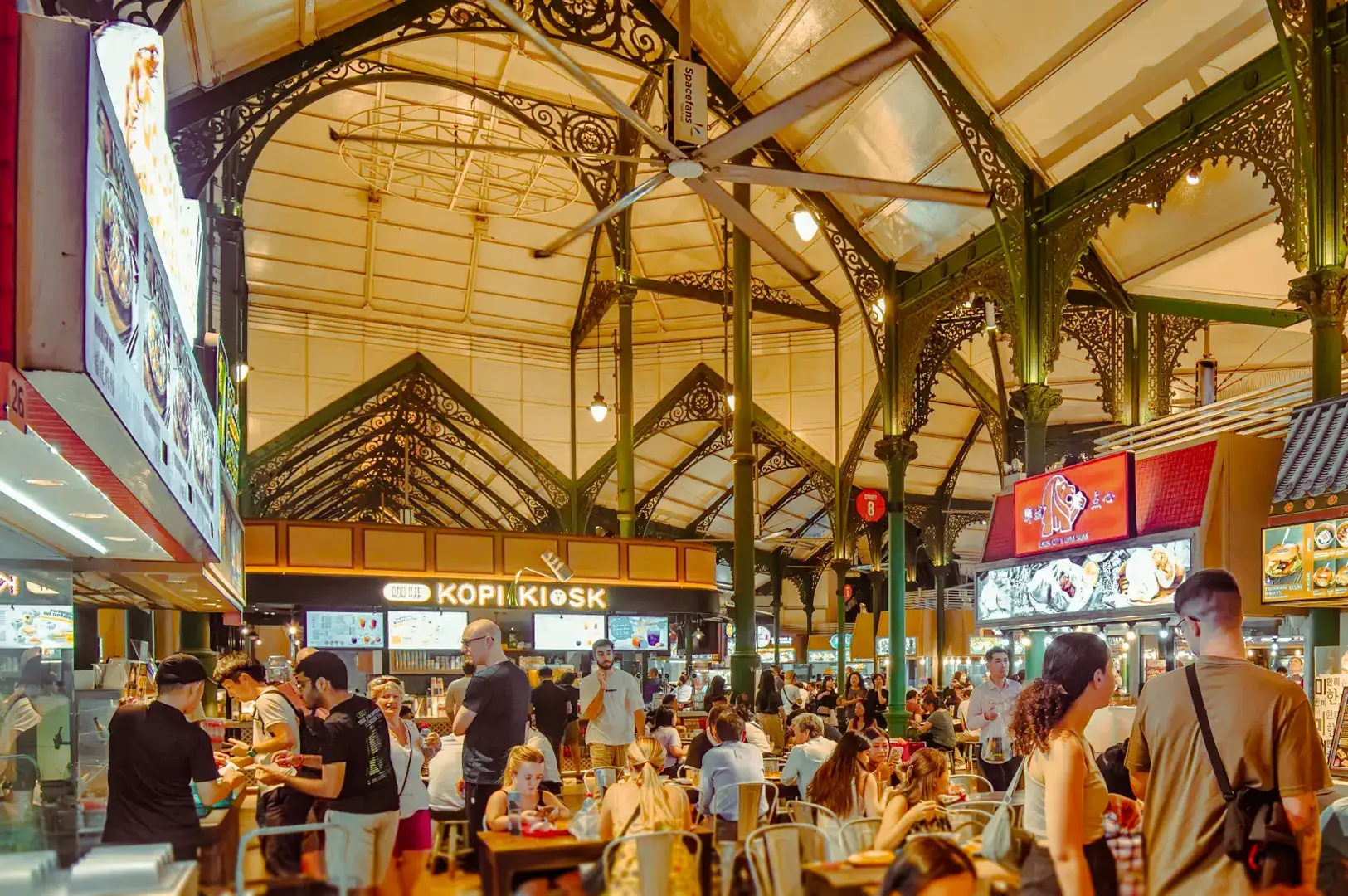 Wide‑angle interior shot of a busy Singapore hawker centre with food stalls, communal seating, and diners under a high metal roof.