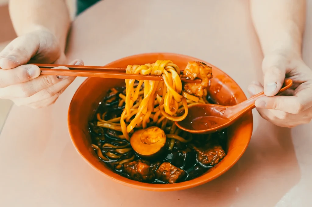 Close‑up, top‑down food shot of a bowl of Singapore lor mee with thick yellow noodles, braised meat, and halved egg in dark gravy, captured from an overhead angle as chopsticks lift noodles above the bowl.