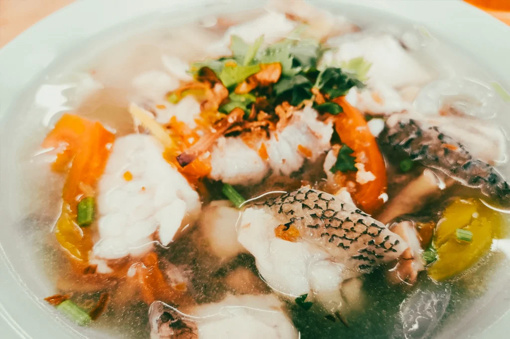 Top-down close-up shot of a clear fish soup with sliced white fish, tomatoes, green herbs, and vegetables in a white bowl, showing delicate textures and light broth.