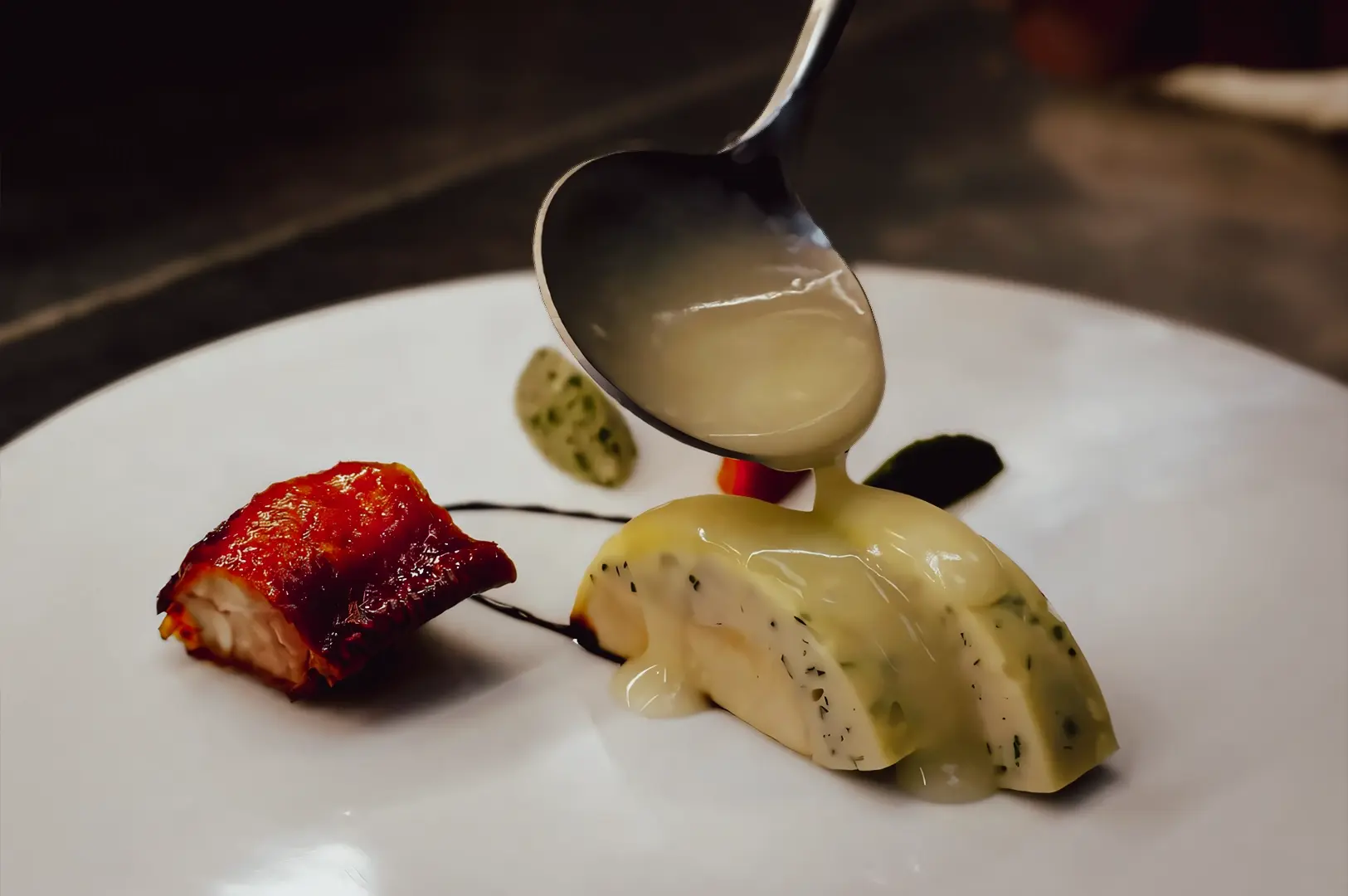 Close-up, slightly top-down shot of a deconstructed Hainanese chicken dish featuring tender poached chicken slices layered with herb-infused elements, aromatic rice components, and a silky ginger-garlic sauce being poured from a spoon onto a minimalist white plate for fine dining presentation.