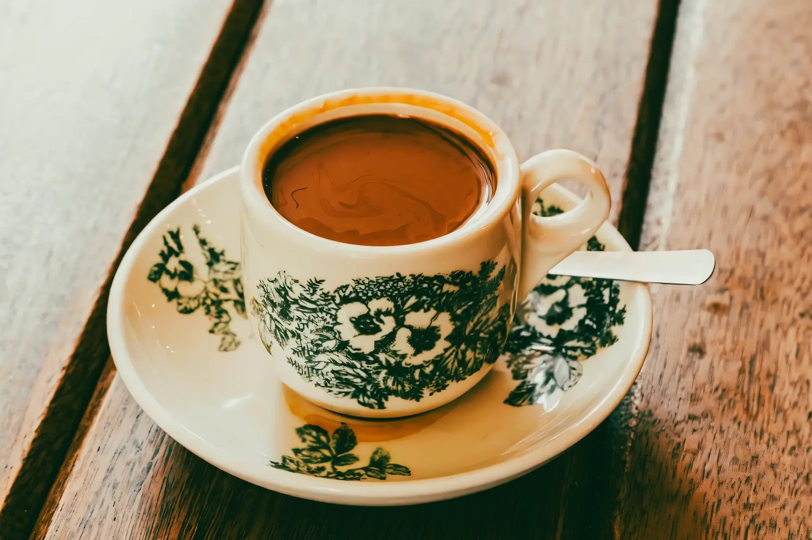 Close-up eye-level shot of a cup of traditional kopi served in a floral-patterned ceramic cup and saucer on a wooden café table.
