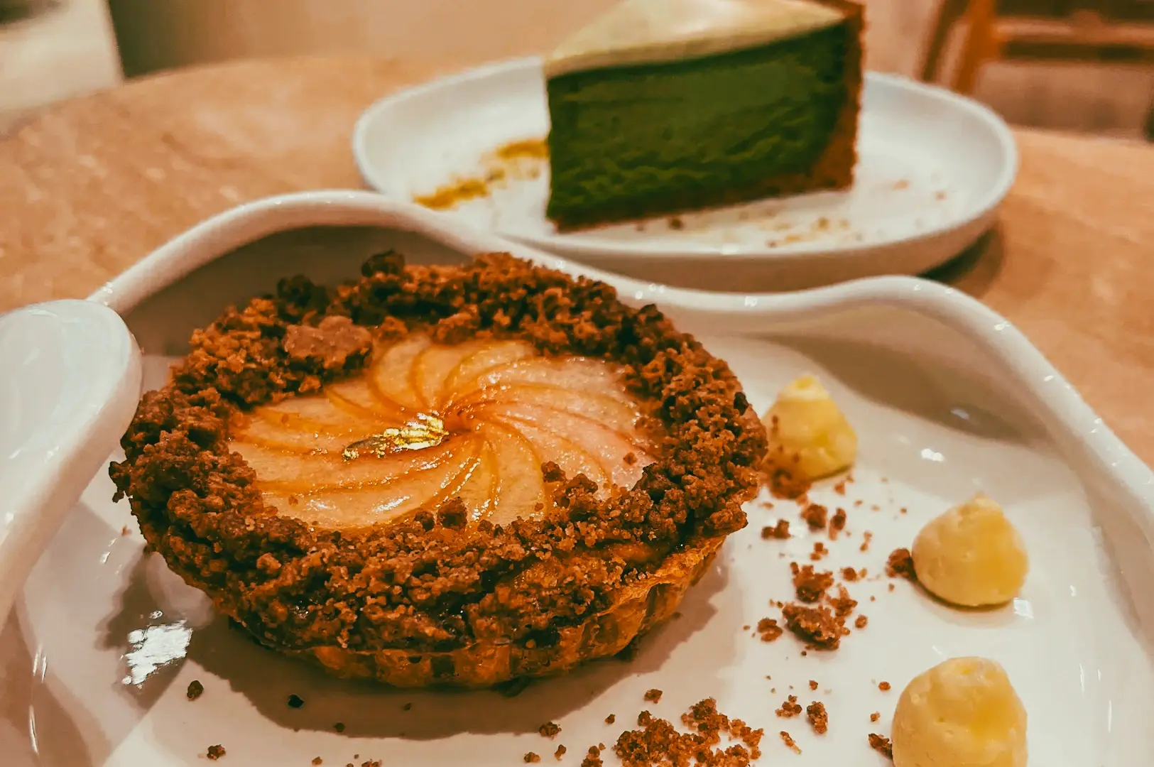 Close-up eye-level shot of a plated café dessert featuring a pear tart with crumb topping and gold leaf, accompanied by small cream dollops, with a slice of green cake on a plate in the background.