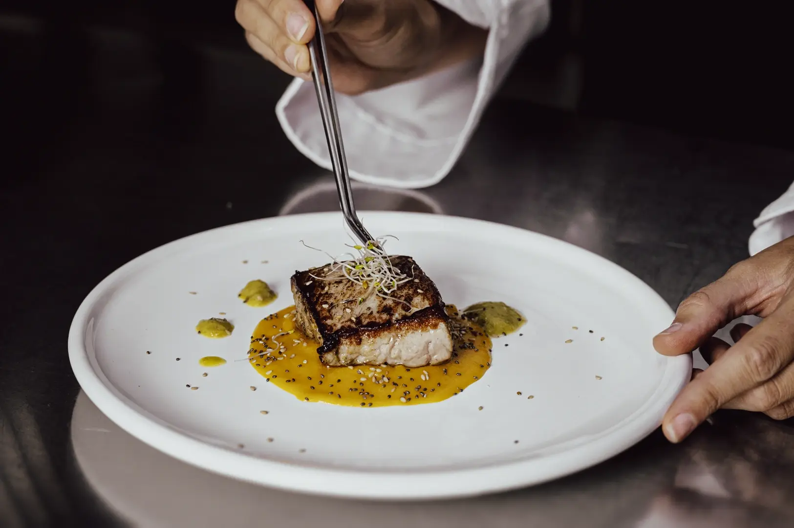 Close-up action shot of a chef plating a gourmet dish with precision tweezers, emphasizing culinary craftsmanship and fine dining preparation.