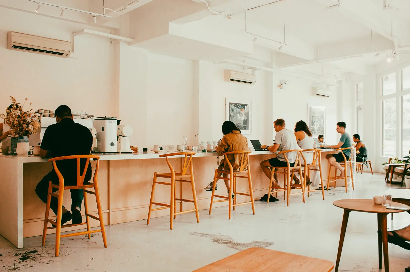 Wide-angle eye-level shot of a bright minimalist café interior with customers seated along a counter on wooden stools and natural light filling the space.