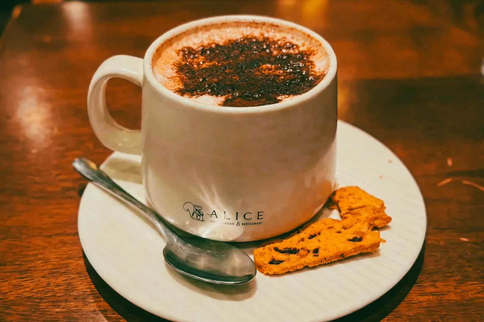 Close-up eye-level shot of a cappuccino topped with cocoa powder, served with a spoon and biscotti on a white saucer at a café table.