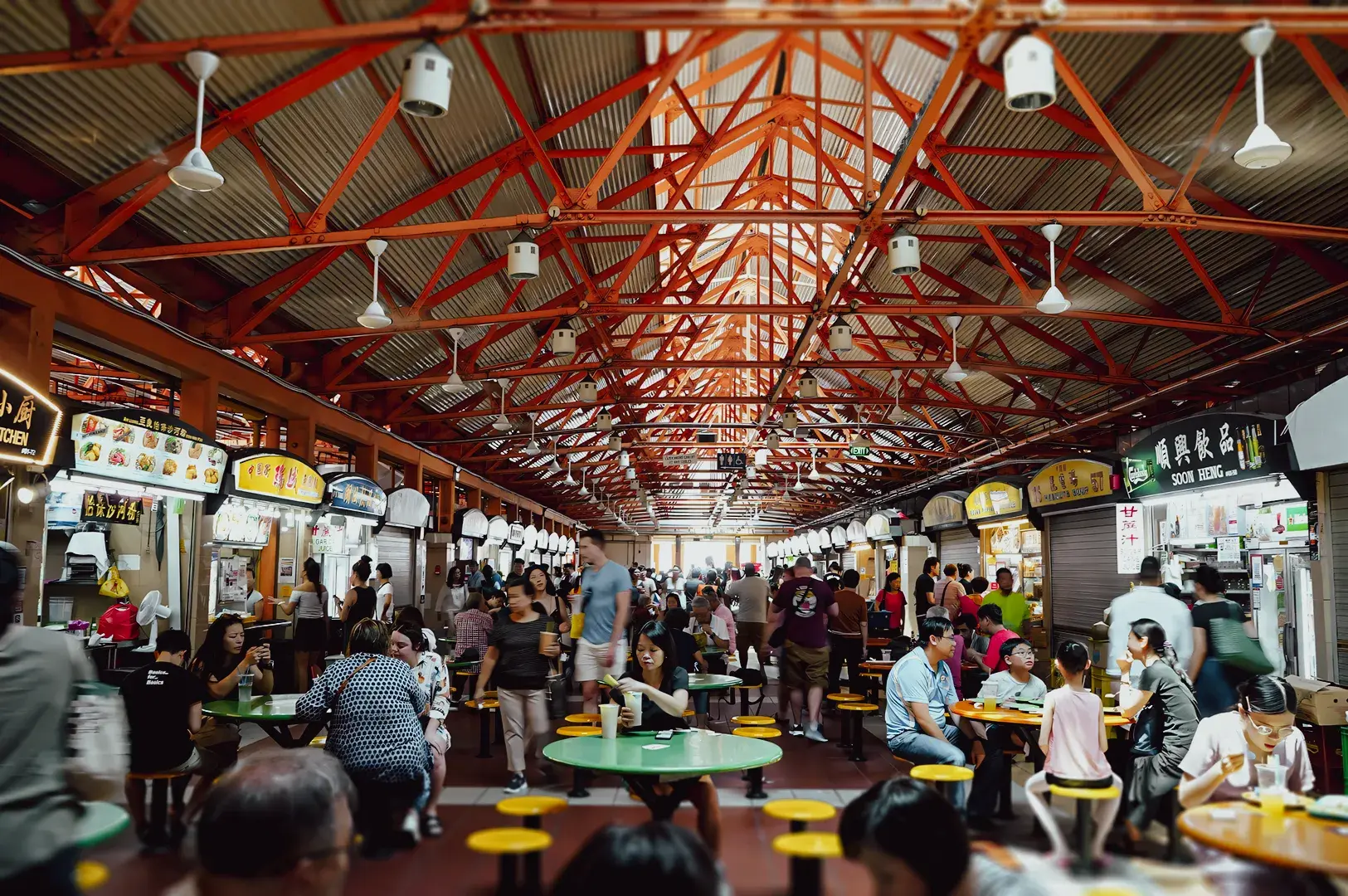 Wide-angle eye-level shot of a busy hawker centre with rows of food stalls, communal seating, and diners enjoying local cuisine, capturing the lively atmosphere of street food dining.