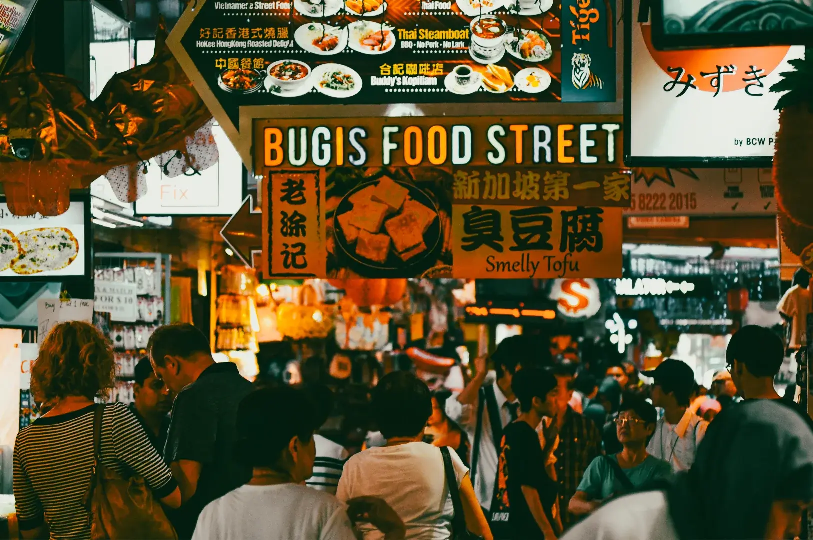 Eye‑level crowd‑level wide shot of Bugis Street food stalls at night, featuring brightly lit signboards advertising street food such as smelly tofu, with dense foot traffic inside the market walkway.
