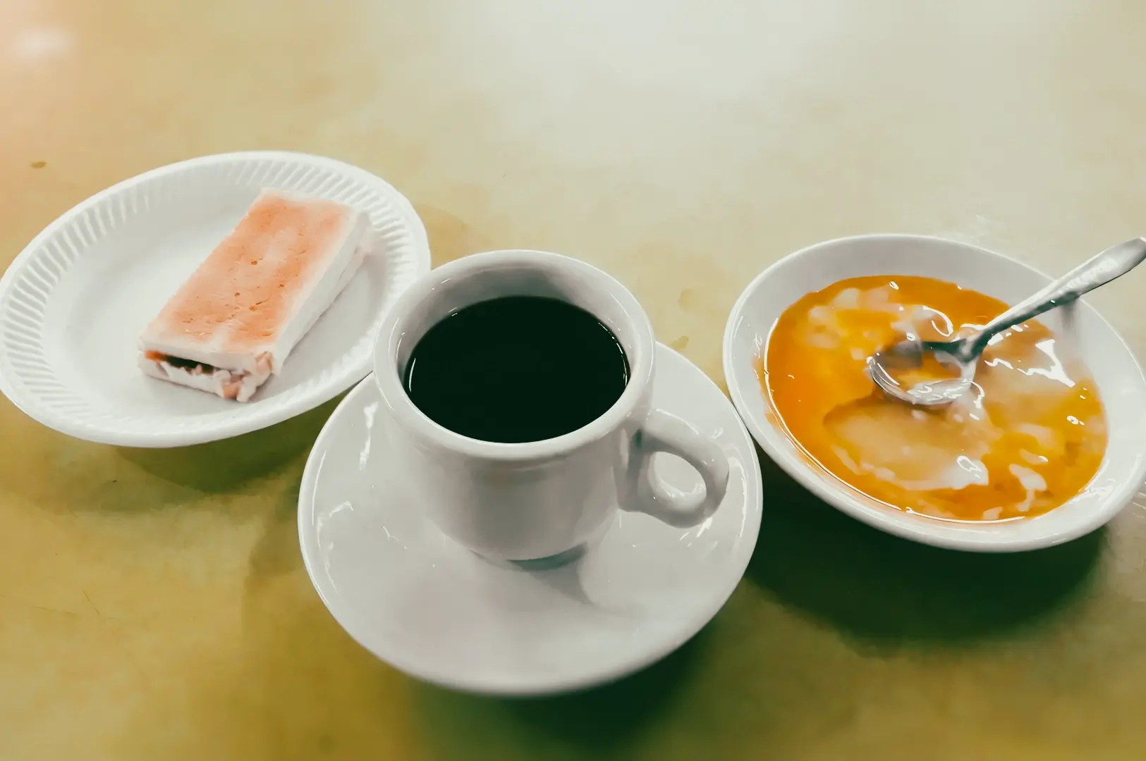 Top-down overhead shot of a traditional coffee set featuring a cup of black coffee, soft-boiled eggs in a bowl, and a kaya toast on a tabletop.