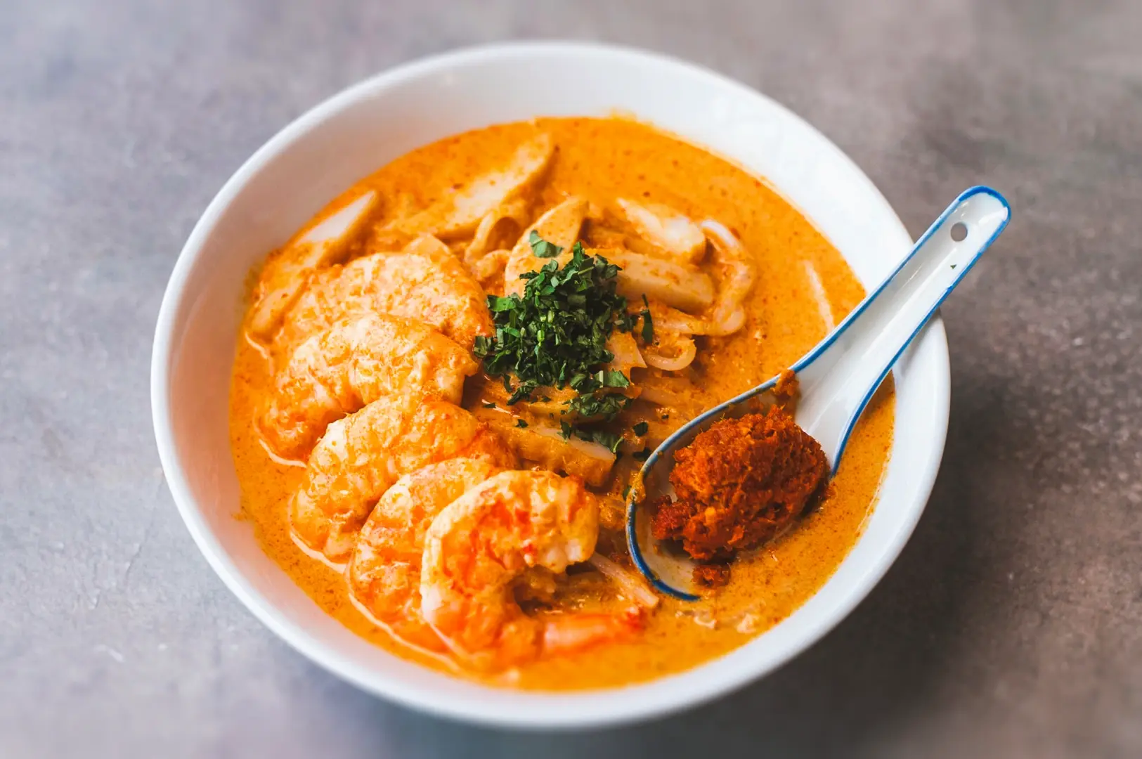 High‑angle close‑up of Singapore Katong Laksa with prawns and fish cake in a creamy spicy coconut curry broth, garnished with herbs.