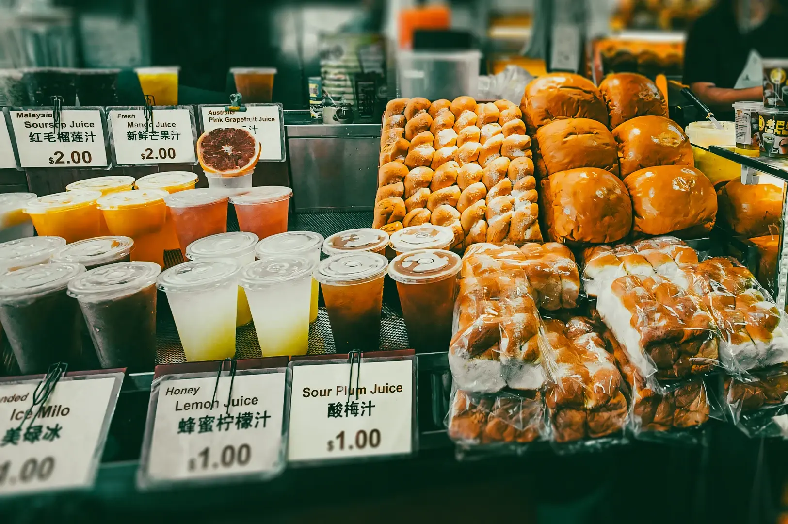 Eye‑level close‑up wide shot of a Bugis Street food stall displaying stacked buns, pastries, and bottled snacks beside rows of takeaway drinks, including sour plum juice and honey lemon juice, with handwritten price tags and a busy hawker stall backdrop.