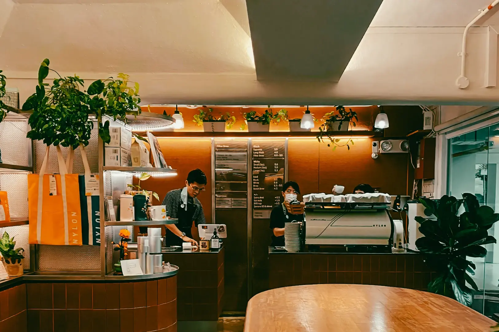 ye-level wide shot of a modern coffee shop counter with baristas preparing drinks behind an espresso machine, surrounded by plants and warm lighting.