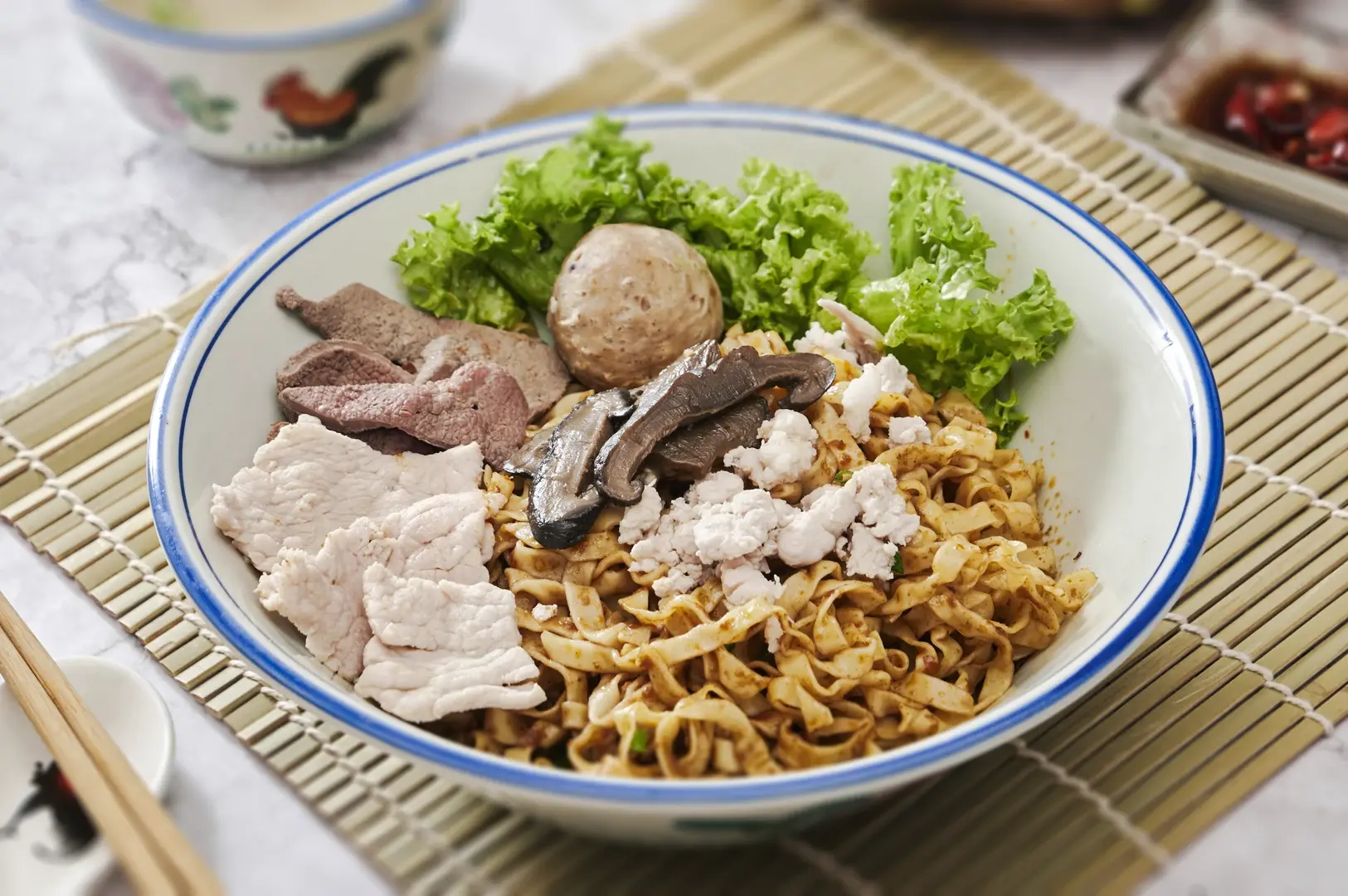 Singapore bak chor mee noodles topped with sliced pork, mushrooms, minced meat, lettuce, and savory sauce in a ceramic bowl.