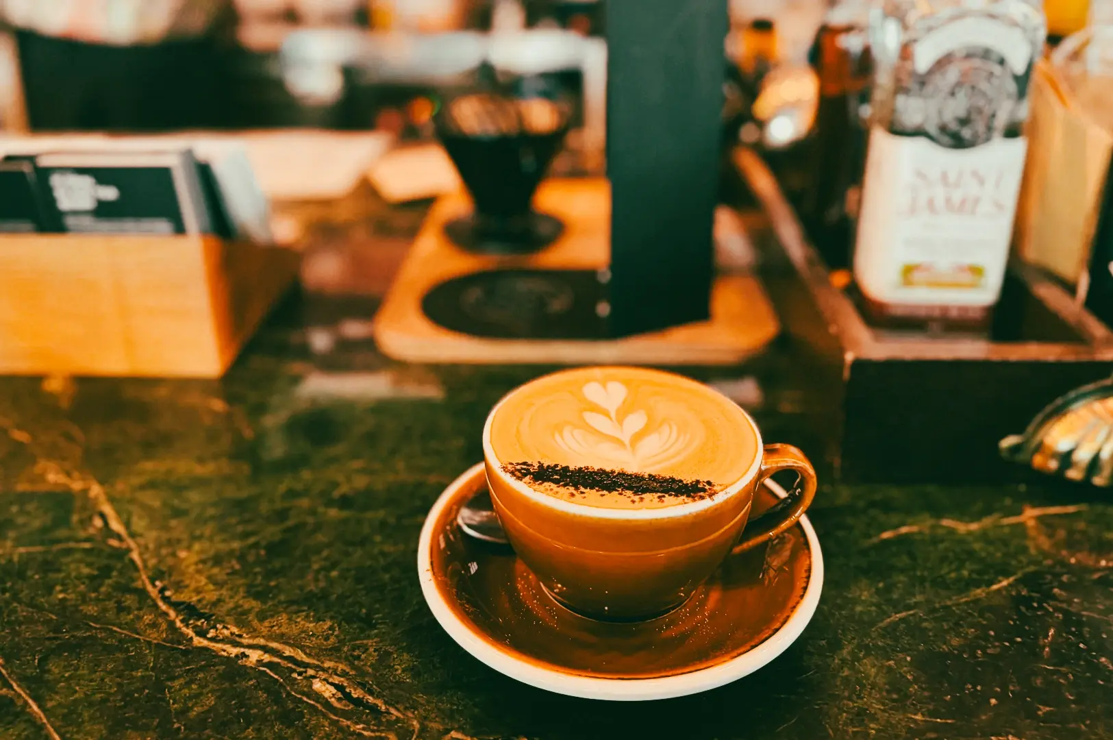 Close-up eye-level shot of a latte with leaf-shaped latte art served in a ceramic cup on a café counter, with coffee equipment blurred in the background.