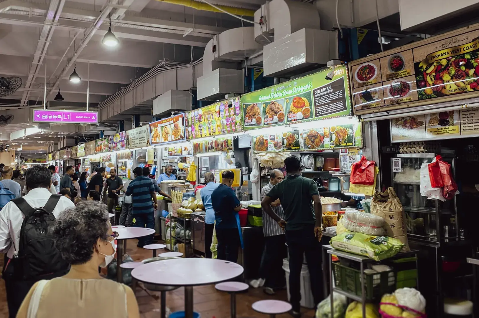 Eye-level wide shot of a bustling indoor food market with multiple food stalls, illuminated menus, and diners lining up, capturing vibrant street food culture.