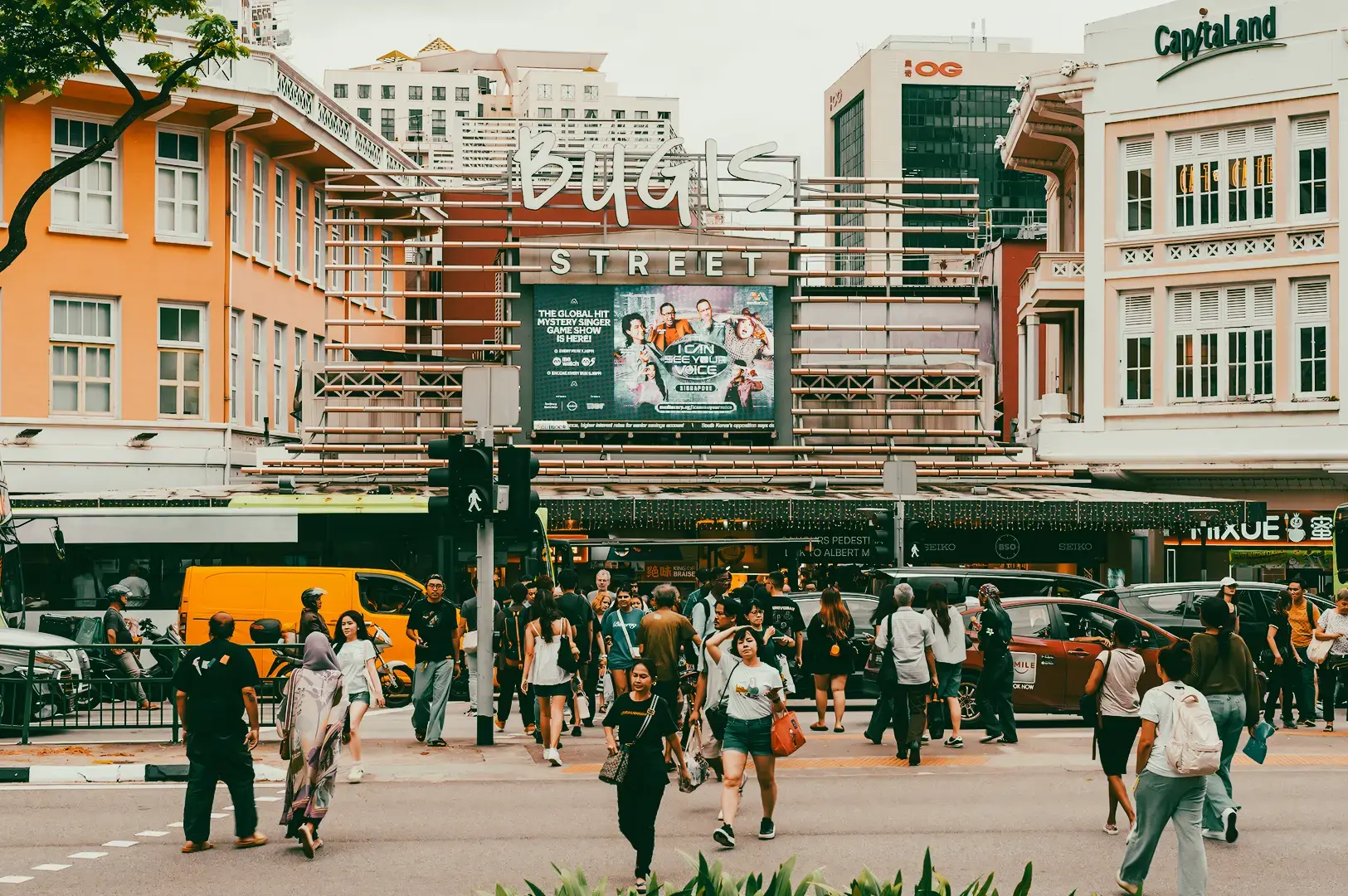 Eye‑level wide‑angle street shot of Bugis Street in Singapore, showing the main entrance signage, surrounding shophouse buildings, and crowds crossing the road in front of the popular shopping and food area.