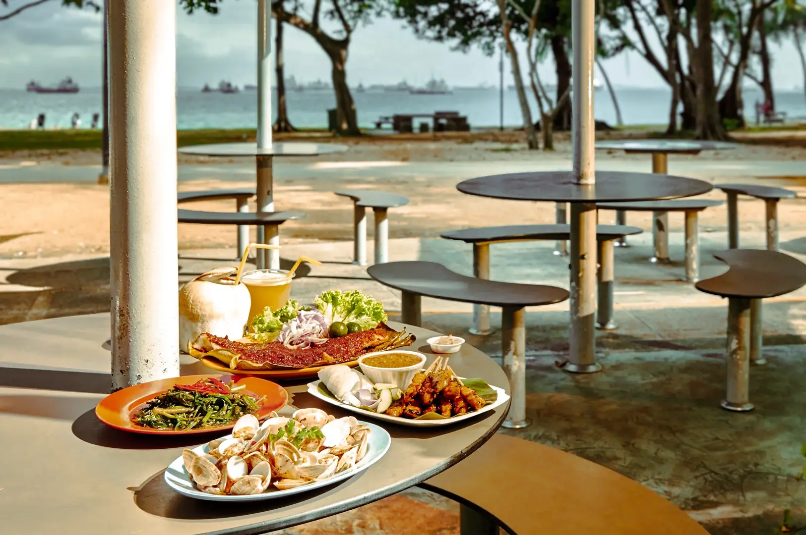 Traditional Singapore hawker food spread with grilled seafood, clams, vegetables, and coconut drink served at an outdoor seaside food court.