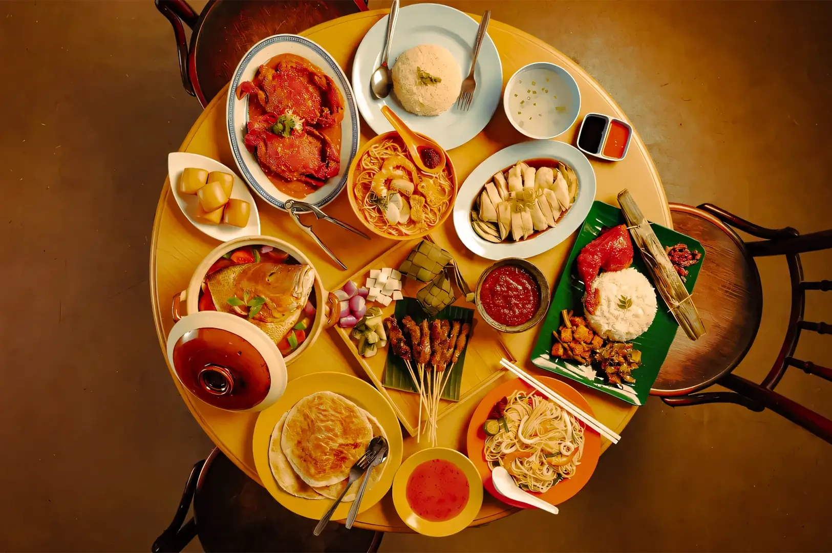 Overhead flat‑lay shot of a traditional Singapore meal spread with multiple hawker dishes, rice plates, noodles, satay, and dipping sauces.