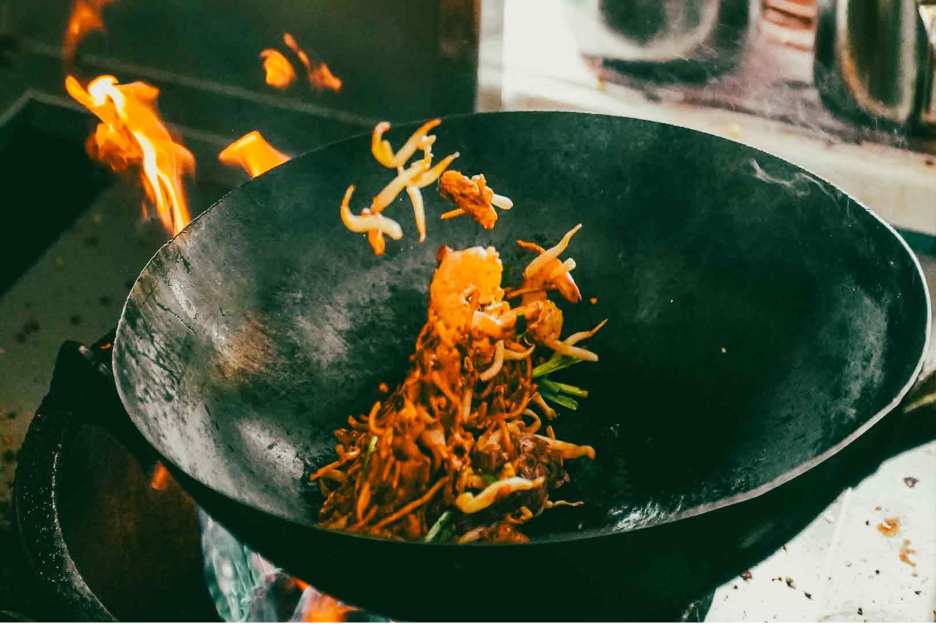 Close-up, slightly low-angle action shot of char kway teow being tossed in a black wok over roaring flames, showcasing intense wok hei and traditional Singapore hawker-style stir-frying.