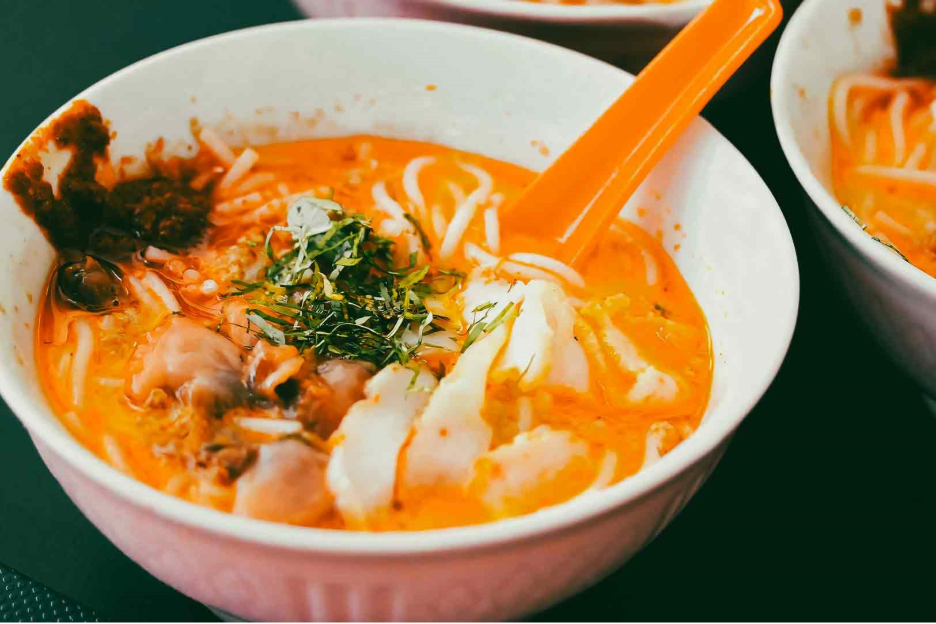 Three-quarter close-up of a bowl of Singapore laksa with rich coconut curry broth, rice noodles, seafood toppings, fresh herbs, and chili, served at a traditional hawker stall.
