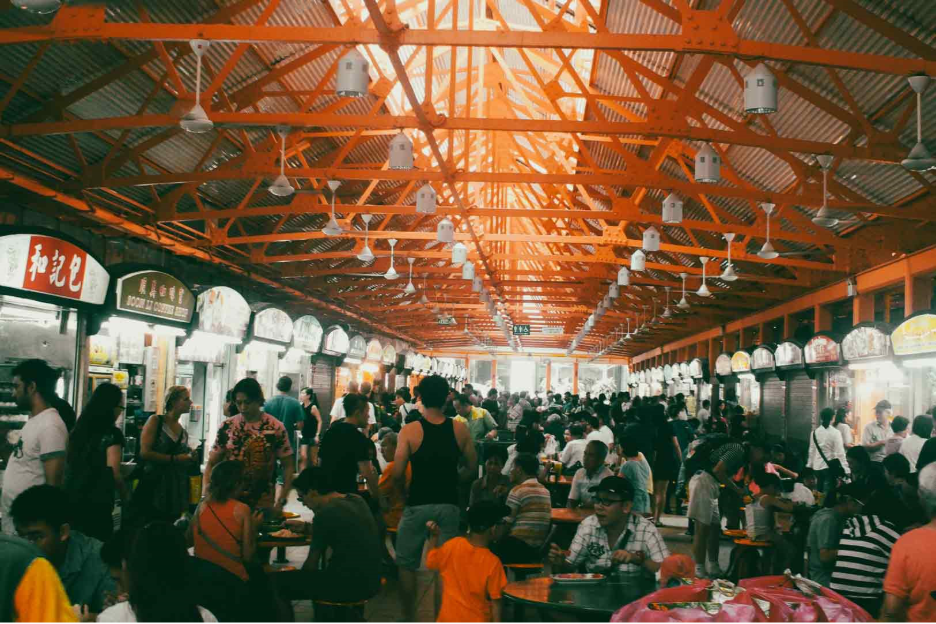 Wide-angle eye-level view of a busy Singapore hawker centre with rows of food stalls, communal seating, exposed orange steel beams, and crowds dining, showcasing local street food culture.