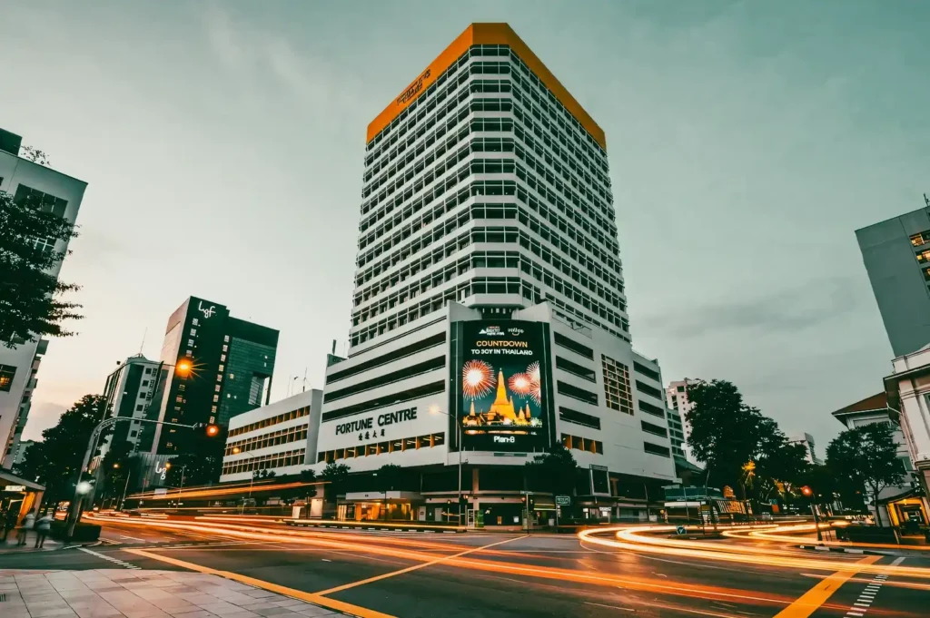 “Wide-angle, low-angle street-level shot of a tall multi-story commercial building at dusk, featuring a white facade with a grid of windows and a contrasting orange rooftop band, surrounded by neighboring urban buildings, with a busy intersection in the foreground captured using long-exposure light trails from passing vehicles, soft ambient evening lighting, and a muted cloudy sky creating a cinematic cityscape atmosphere.