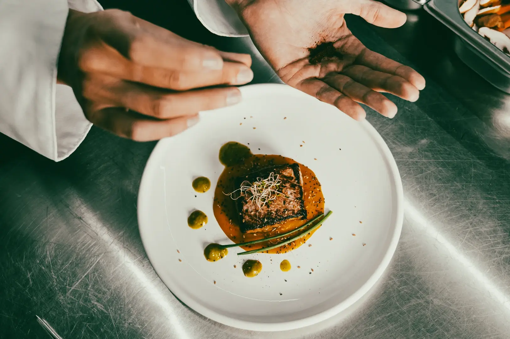 Chef plating a signature fine dining dish with sauce and garnish at a restaurant, captured from an overhead top-down angle on stainless steel work surface.