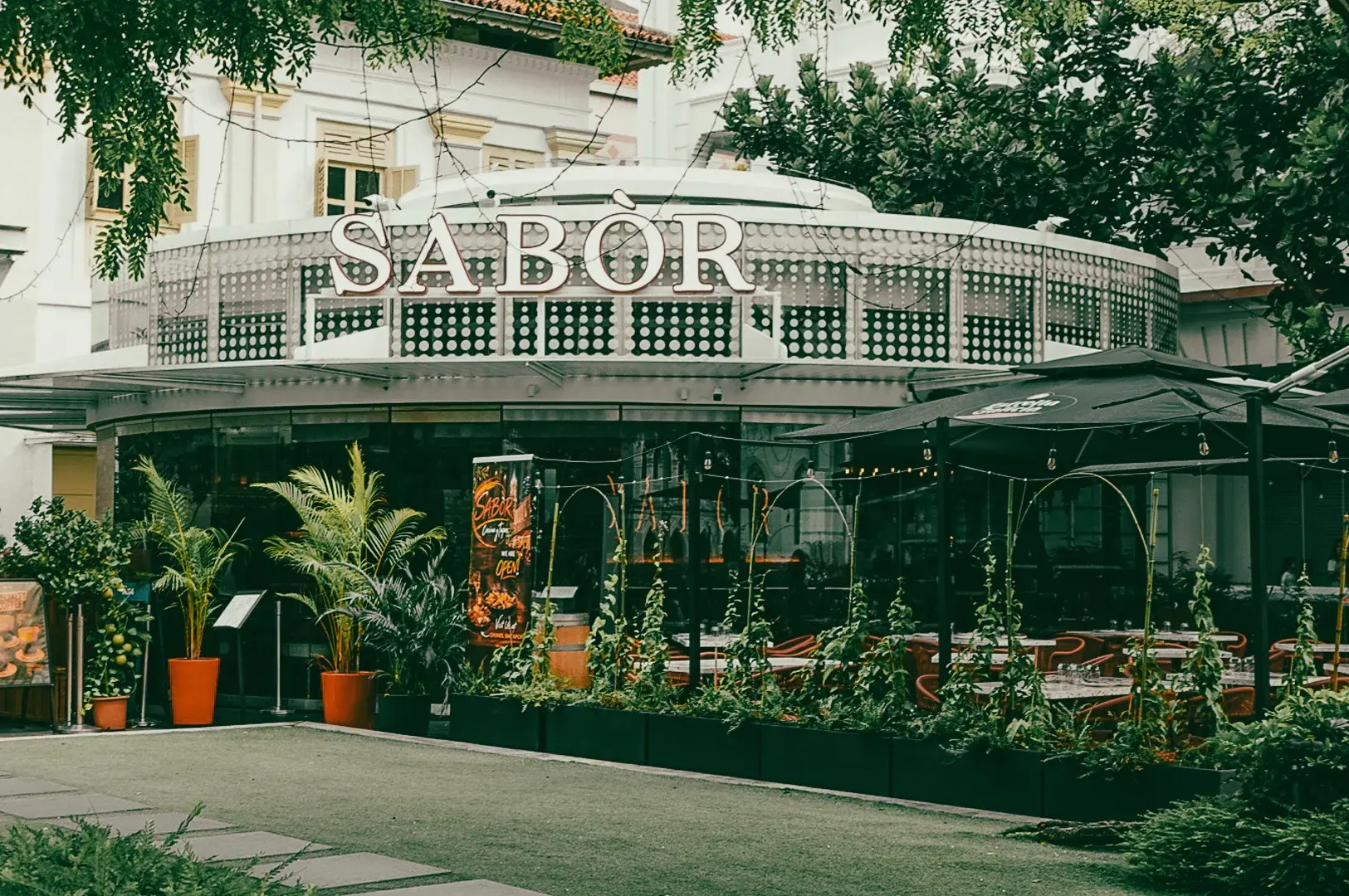 Wide-angle exterior shot of Sabor restaurant at CHIJMES Singapore with alfresco dining area, greenery, and heritage building facade near City Hall