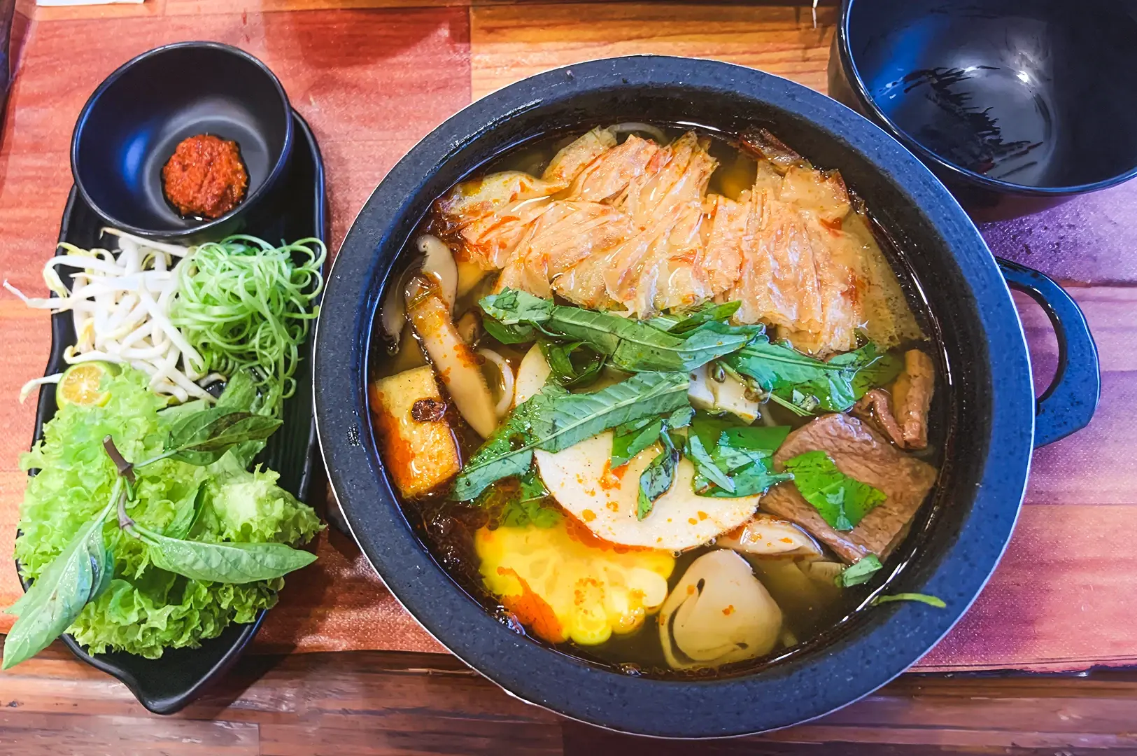 Top-down close-up view of a bowl of vegetable soup with napa cabbage, leafy greens, mushrooms, tofu, and sliced plant-based ingredients in broth, served in a black pot on a wooden table, with side dishes including bean sprouts, lettuce, herbs, calamansi, and a small dish of chili paste arranged on a rectangular plate under bright indoor lighting.
