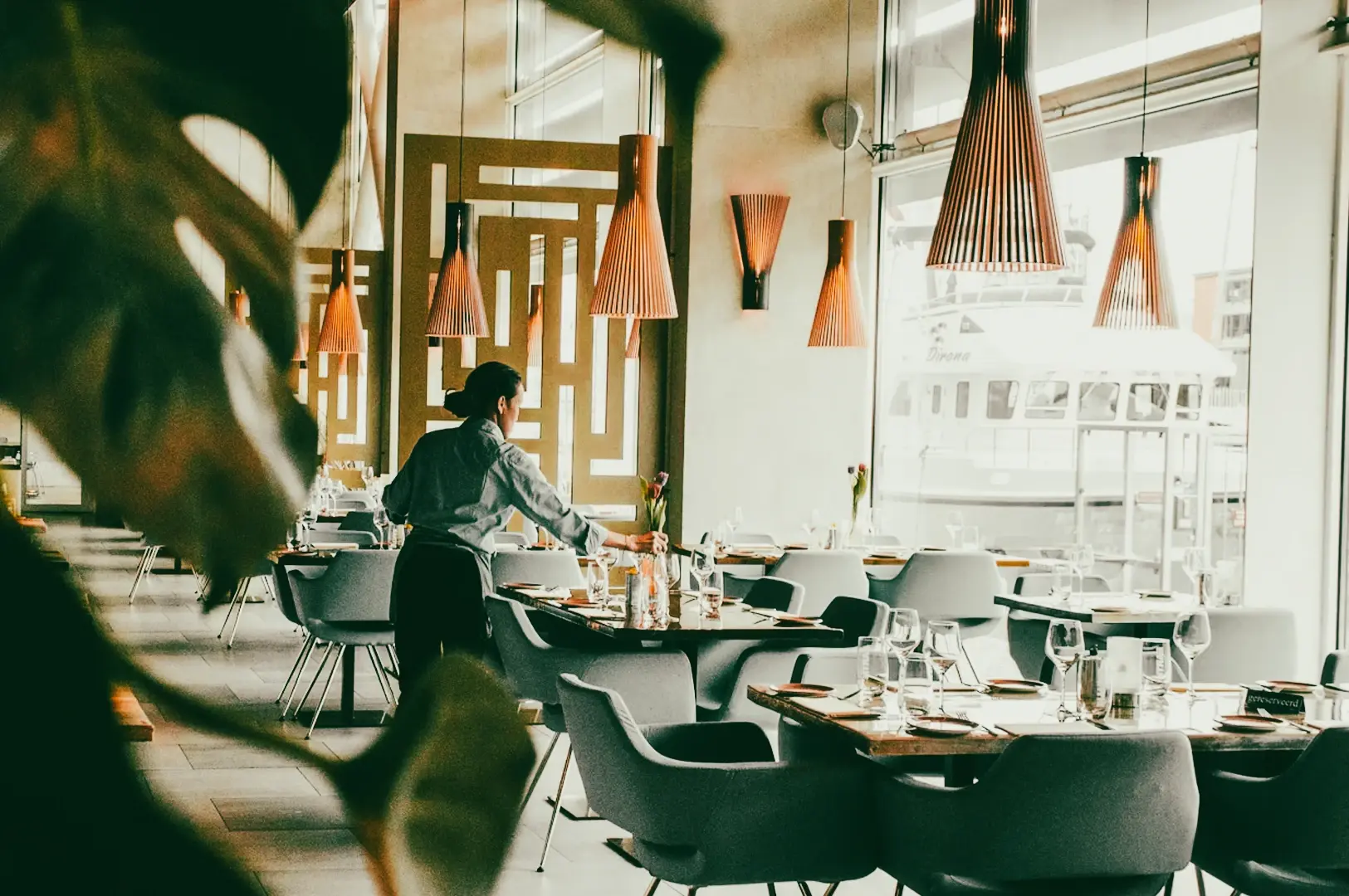 Modern restaurant interior with contemporary chairs, pendant lighting, and floor-to-ceiling windows, photographed from a wide-angle perspective during table setup.