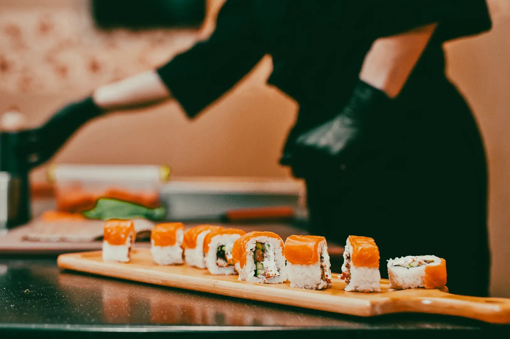 Sushi rolls lined up on a wooden board at a Japanese restaurant, captured from a low side-angle perspective with chef workspace in the background.