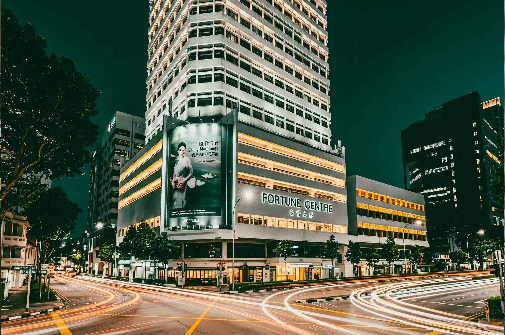 Low-angle wide-angle night view of Fortune Centre building at a city intersection, showing a tall multi-story structure with illuminated horizontal bands, large billboard advertisement on the facade, and ‘FORTUNE CENTRE’ signage above the entrance, with surrounding office buildings, trees, streetlights, and long-exposure light trails from passing vehicles on the road.