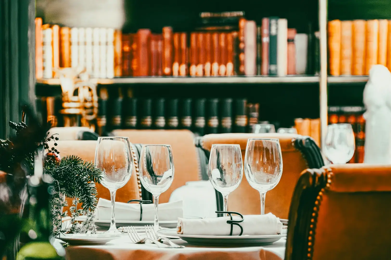 Elegant table setting inside a restaurant with wine glasses, napkins, and leather chairs, photographed at eye-level angle with bookshelf-lined dining room in the background.