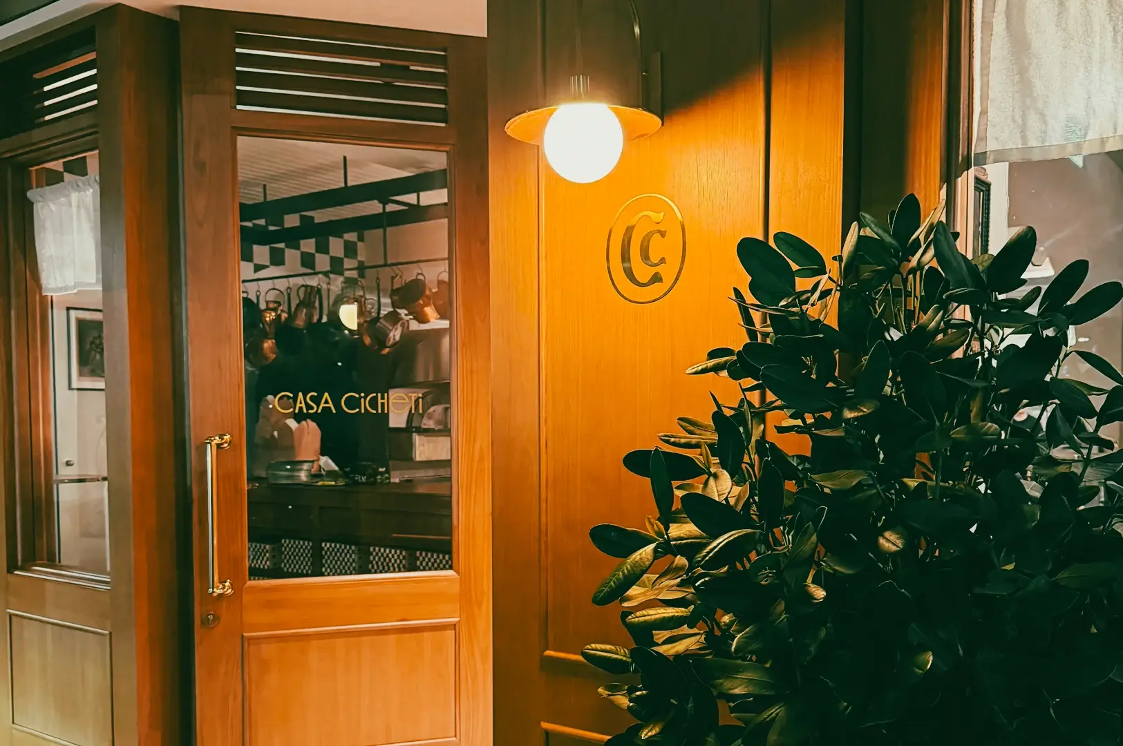 Eye-level medium shot of Casa Cicheti entrance with wooden door, warm lighting, and indoor kitchen view, showcasing cozy Italian restaurant ambiance in Singapore