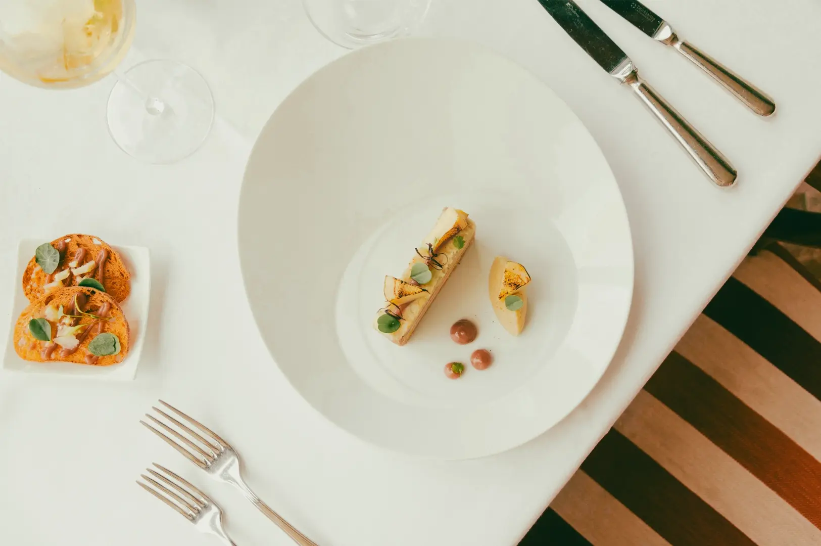 Overhead flat lay shot of a fine dining plated appetizer with foie gras terrine, pear slices, microgreens, and sauce dots on a white plate, styled on a minimalist restaurant table setting.