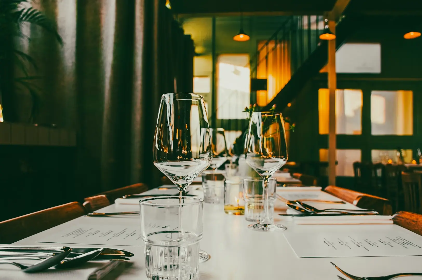 Eye-level, shallow depth-of-field shot of a long fine dining table set with clear wine glasses, water tumblers, neatly arranged cutlery, and printed menus atop a white tabletop; warm ambient lighting reflects through glassware, while upholstered booth seating, soft greenery along the wall, and warmly lit windows recede into the background, highlighting symmetry, refined textures, and an upscale Singapore restaurant interior.