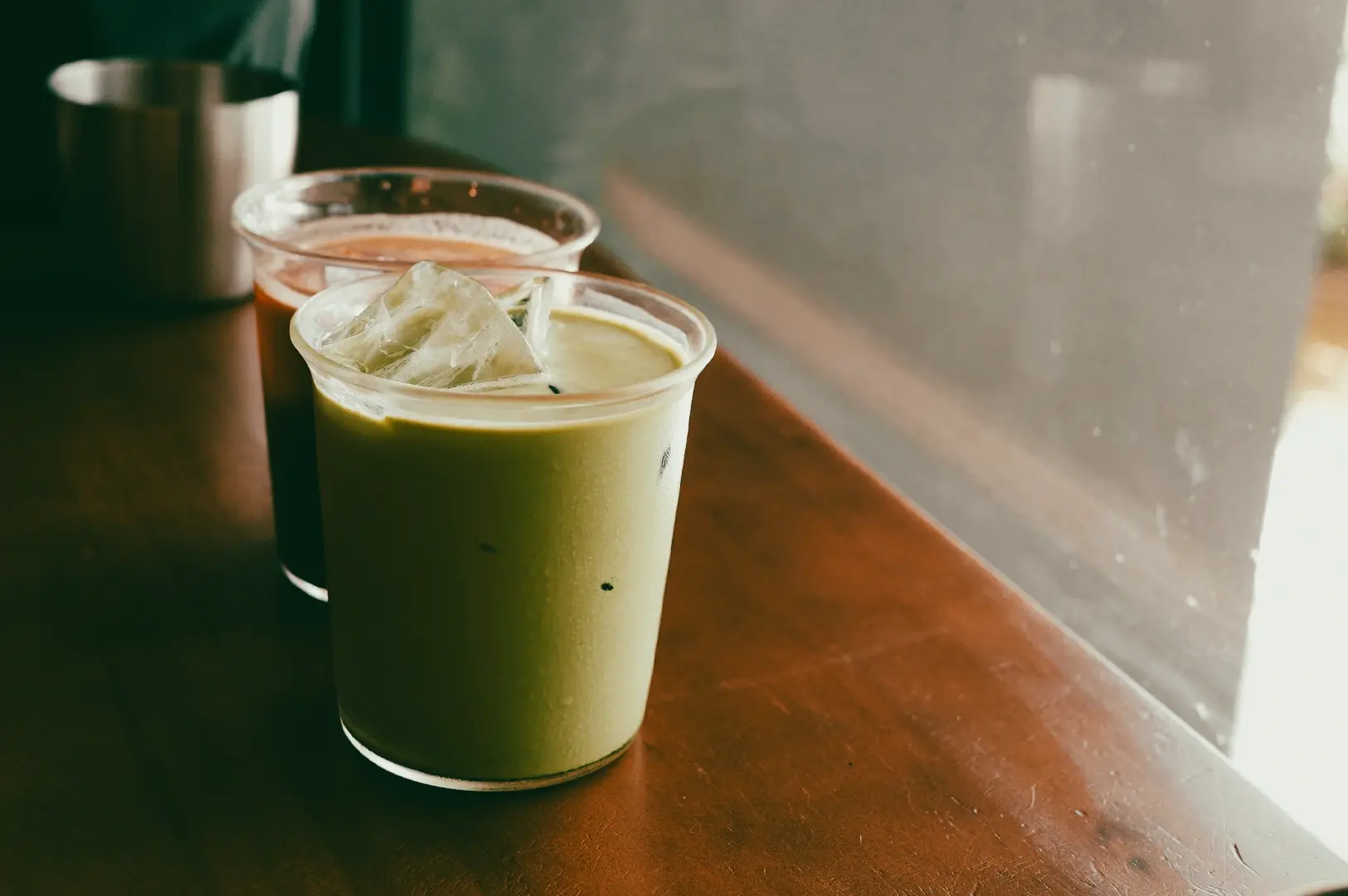 Side-angle close-up shot of iced matcha latte and iced coffee by a window in a Singapore cafe, representing refreshing specialty drinks and modern cafe culture